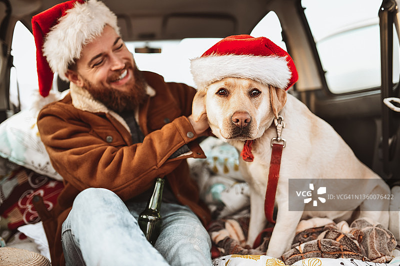 一名大胡子男子在小货车上给他的宠物狗戴圣诞帽时面带微笑图片素材