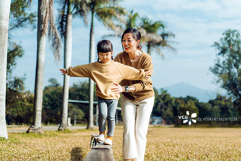 在一个阳光明媚的日子里，年轻的亚洲妈妈和小女儿在自然公园里度过了快乐的时光。母亲扶着小女儿，双臂伸展在木板上保持平衡。家庭生活方式，关爱理念图片素材