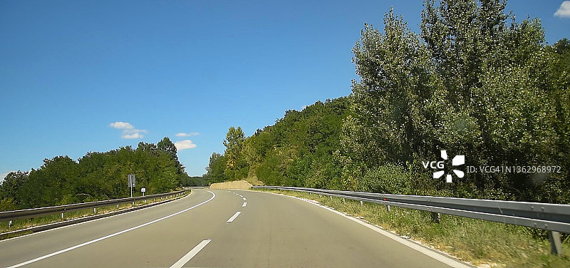 在阳光明媚的夏日，宽阔的道路周围是绿色的田野和树木。图片素材