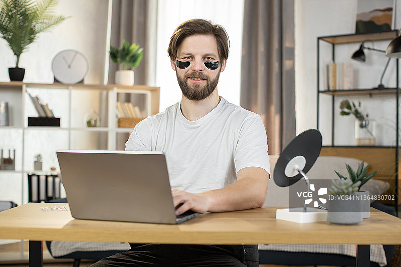 白种人，戴着黑色的胶原蛋白眼罩，坐在家里的桌子上用笔记本电脑工作图片素材