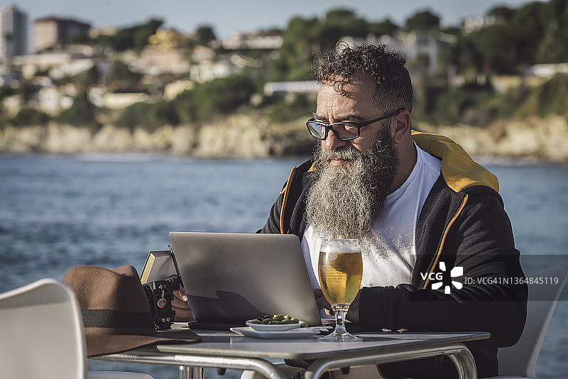留着长胡子的Homabre坐在海边的一家咖啡馆里，一边用他的笔记本电脑在线工作。图片素材