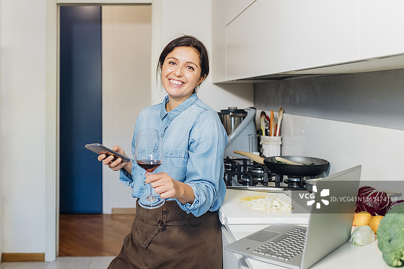 女人拿着酒杯和智能手机靠在厨房的柜台上在家图片素材