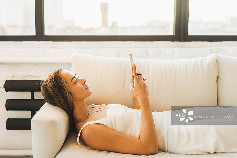 无聊的女人躺在沙发上玩手机。把时间浪费在网络上。拖延症与设备图片素材