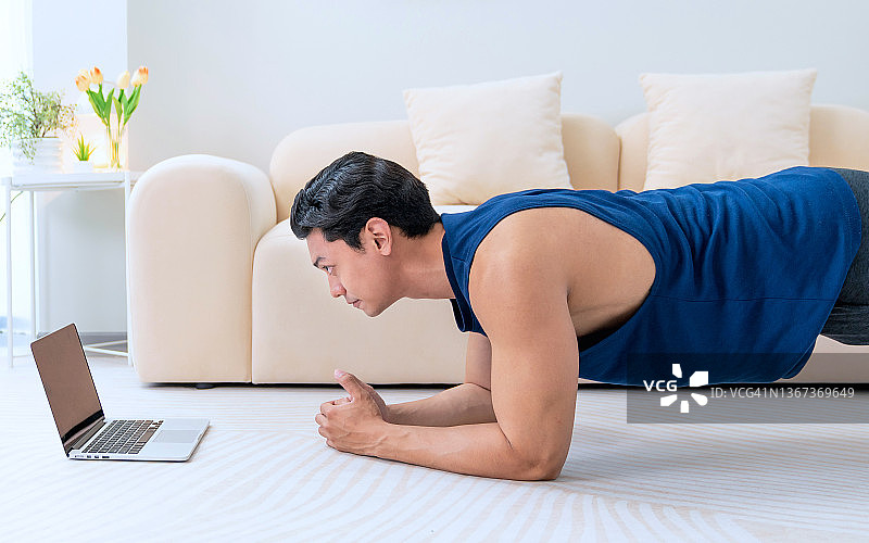 男人在家训练。运动男子做瑜伽平板，同时看在线教程的笔记本电脑。在客厅里锻炼。图片素材