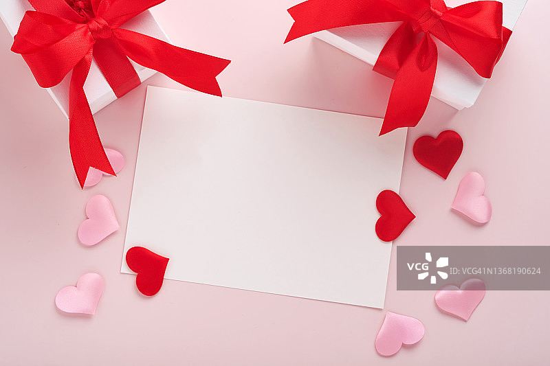白色的礼盒，红色的丝带和小的红色装饰心在粉红色的背景上。顶部视图与复制空间。情人节或婚礼的浪漫概念。节日的作文。模拟。图片素材
