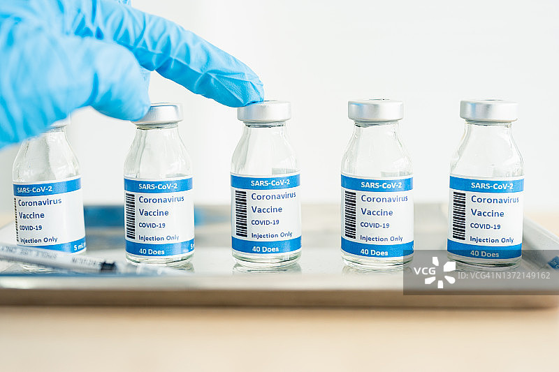 冠状病毒疫苗瓶在实验室的长凳上。背景中，一名卫生保健专业人员正在整理一盘COVID-19疫苗的小瓶和注射器图片素材