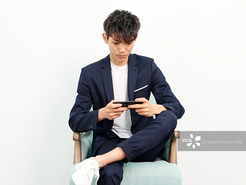 英俊的中国年轻人的肖像，在深蓝色的休闲西装坐在扶手椅和摆姿势的白墙背景。两腿交叉看着手机，年轻人的生活方式。图片素材