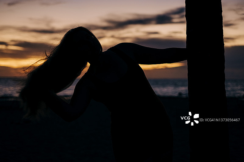 一位妇女斜靠在一棵棕榈树上，垂下头发看日落图片素材