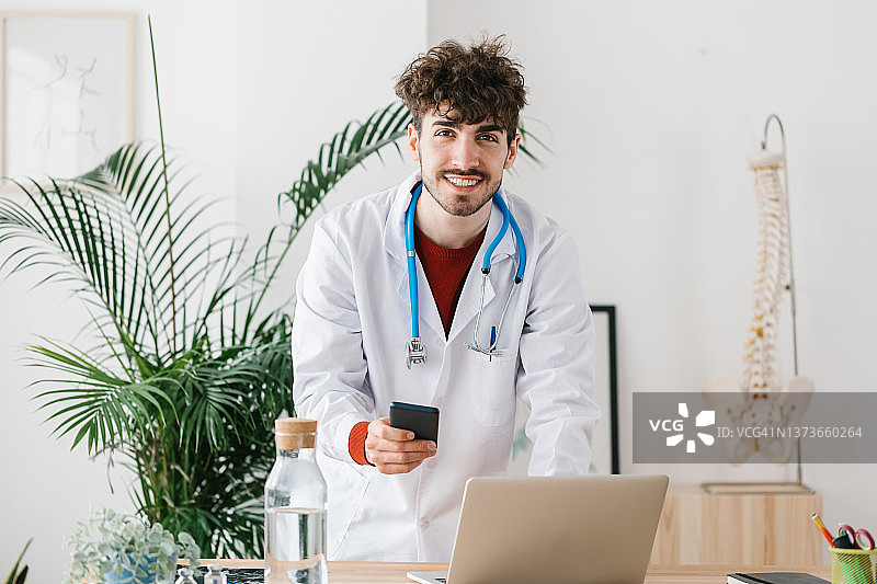 自信的年轻西班牙裔医生使用移动电话，在书桌上的笔记本电脑在医疗办公室图片素材