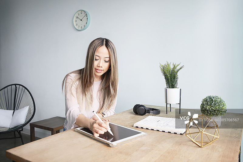一位留着长发的亚洲千禧一代女性穿着休闲服装，在灯光明亮的阁楼空间里，在办公桌前工作时，正在使用数字平板电脑。图片素材