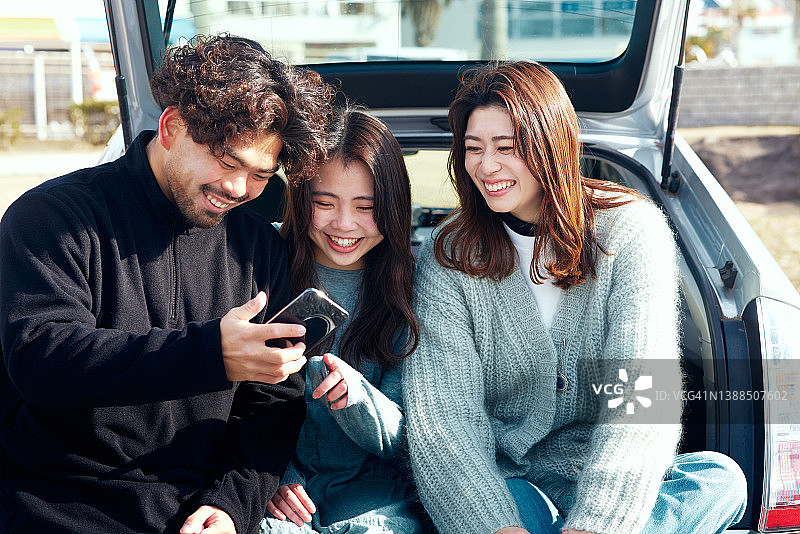 三个朋友坐在车后面，看着他们的智能手机。图片素材