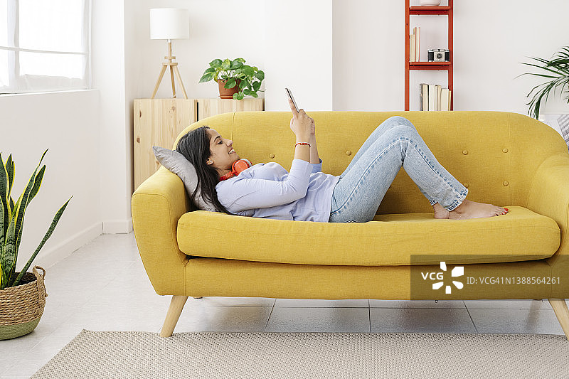 放松的女人微笑，而使用她的手机躺在家里的沙发上。图片素材