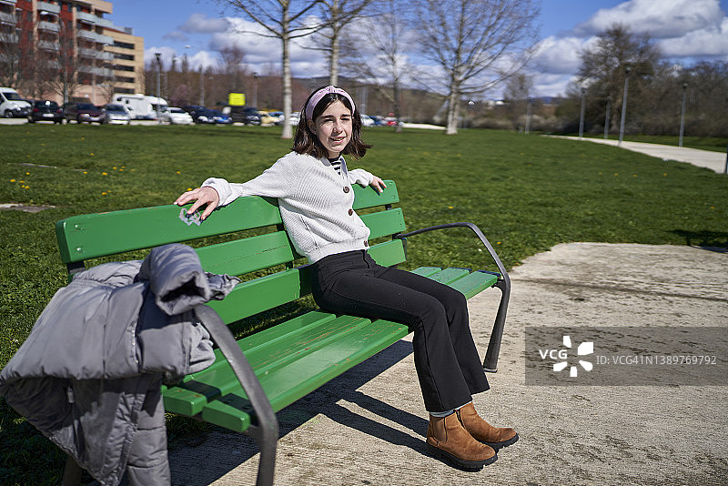美丽的13岁女孩坐在公园的长椅上，在一个阳光明媚的春天，穿着休闲的衣服，看着镜头微笑。图片素材