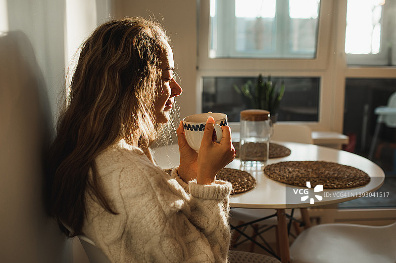 年轻漂亮的女人在家里享受早晨的咖啡。厨房里阳光明媚。图片素材