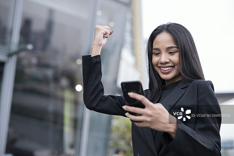 低角度的看法，年轻的商业女性看着智能手机和欢呼与胜利的表情在外面的商业办公室。图片素材