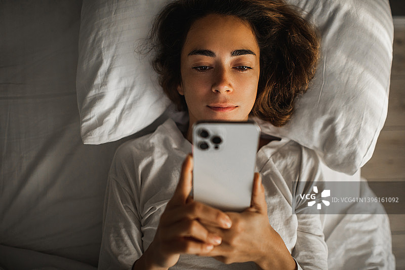 沉迷于网络和小玩意。在智能手机上浪费时间。女人躺在床上拿着电话图片素材