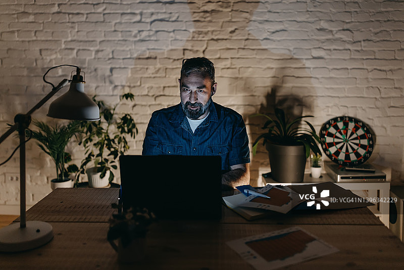成熟的男人穿着休闲西装坐在家庭办公室的桌子上，在笔记本电脑上工作图片素材