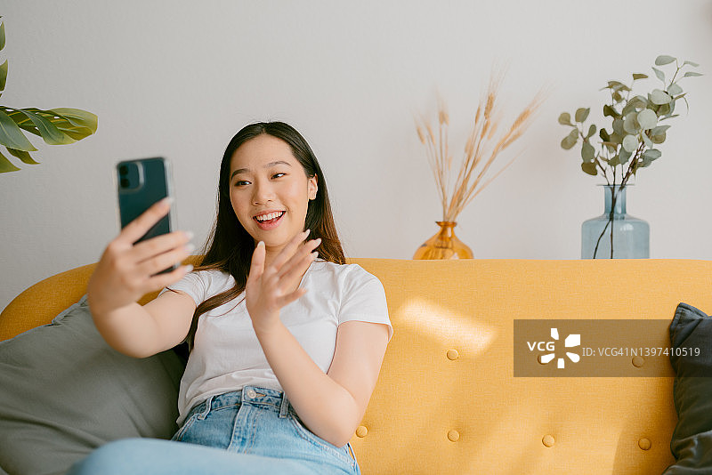 年轻的亚洲女性坐在家里的沙发上喝着咖啡，一边用手机打视频电话，一边微笑着挥手。图片素材