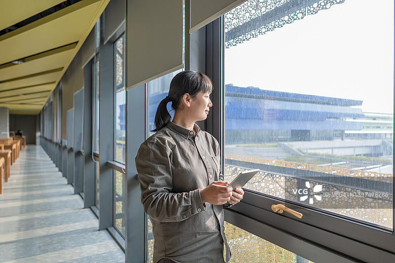 一位亚洲白领女性拿着平板电脑站在窗前向外望去图片素材
