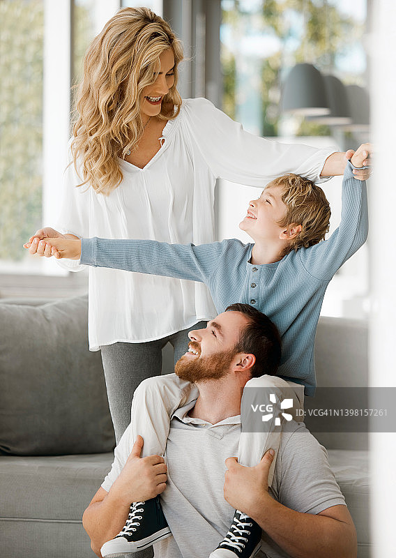快乐的白人家庭关系在家里。兴奋的小男孩坐在爸爸的肩膀上，牵着妈妈的手，假装飞起来，玩得很开心。慈爱的父母和儿子一起放松图片素材