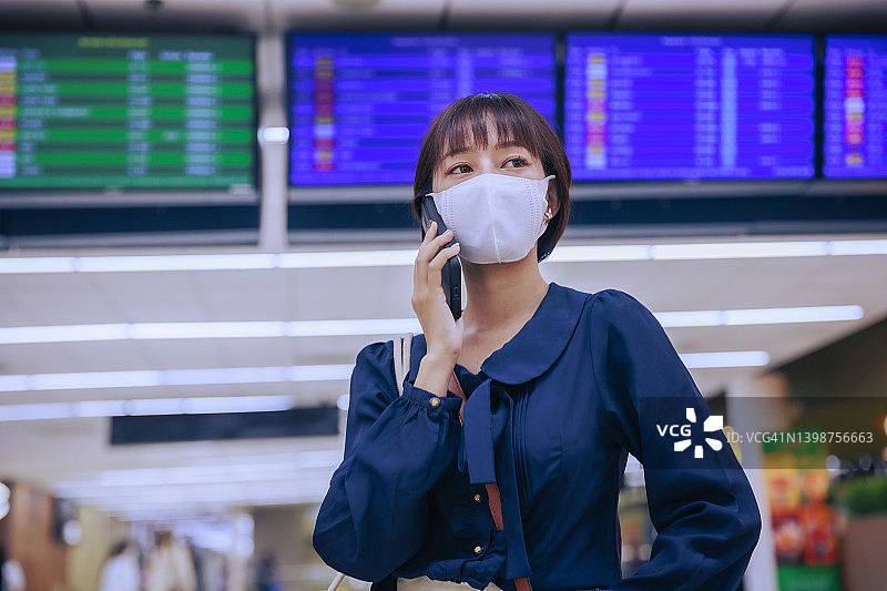 年轻的亚洲女商人戴着口罩站在机场出境区讲手机。商务旅行的概念。图片素材