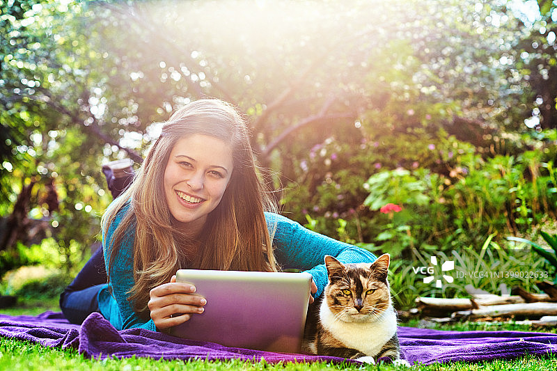 美丽的年轻女子和她的宠物猫躺在户外花园的毯子上，用一个数字平板电脑图片素材