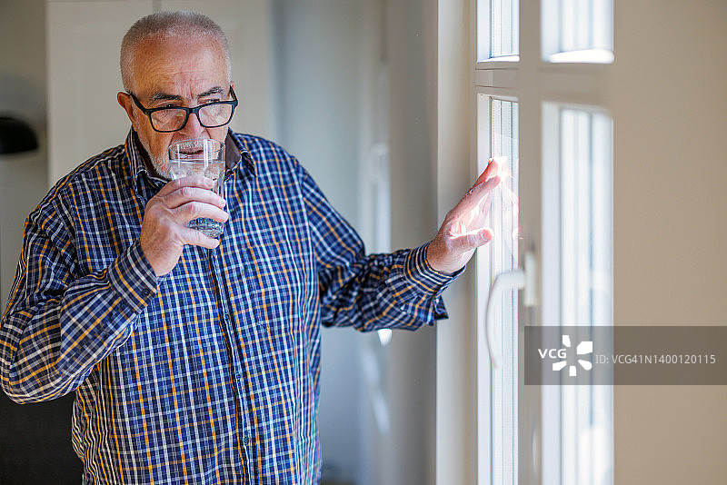 老人喝着水，坐在窗边享受，看着阳光明媚的一天开始图片素材