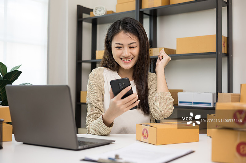 快乐年轻的亚洲女企业家在智能手机上检查产品。商务女性工作中小企业在线在家。请检查包裹上的地址。小企业主开始自由职业的概念。图片素材