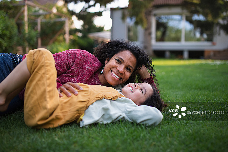 多种族妇女和她的女儿躺在花园里的草地上，享受夏天的时光。图片素材