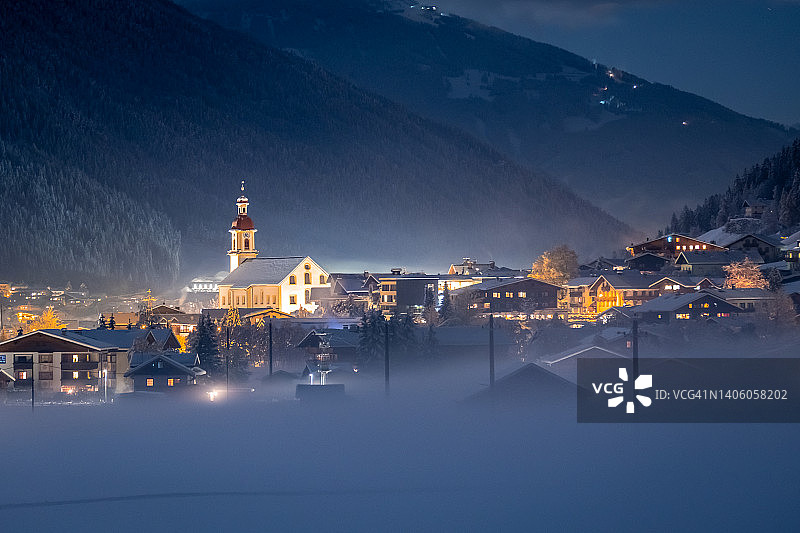 阿尔卑斯山的冬季景观。在一个寒冷的冬天的晚上，在一个寒冷的雾中，一个舒适的村庄。美丽的照明高山房屋和教堂图片素材