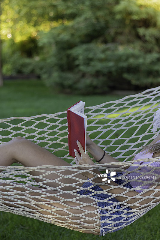 从垂直角度看，不可辨认的年轻女子在夏天的服装阅读一本红色的书与副本的空间在花园图片素材
