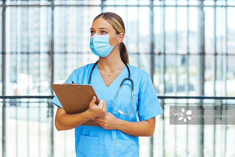 白人女医生或护士护理人员身穿蓝色制服，戴着抗冠状病毒Covid - 19防护口罩，拿着剪贴板。图片素材