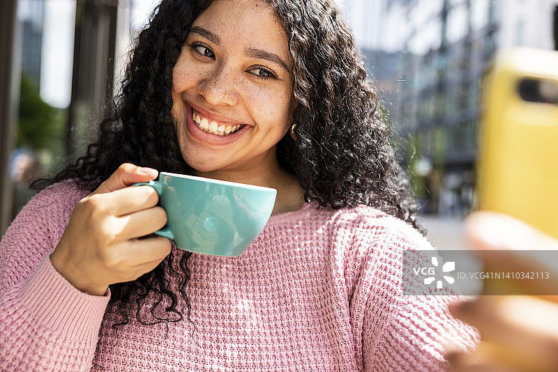 阳光明媚的日子里，快乐的女人拿着咖啡杯用智能手机自拍图片素材