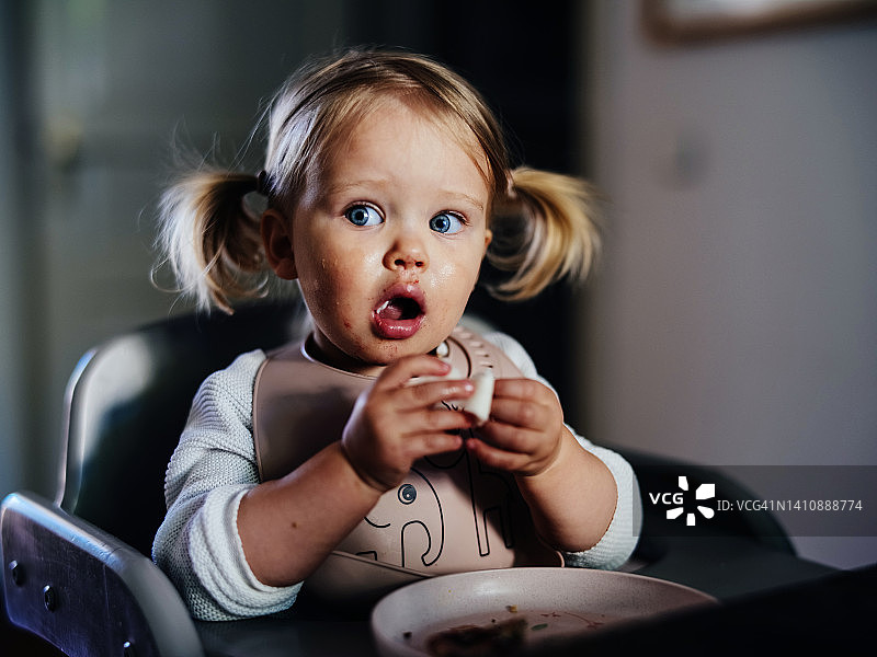 惊奇可爱的小女孩扎着小辫在家里的餐桌上吃饭。图片素材