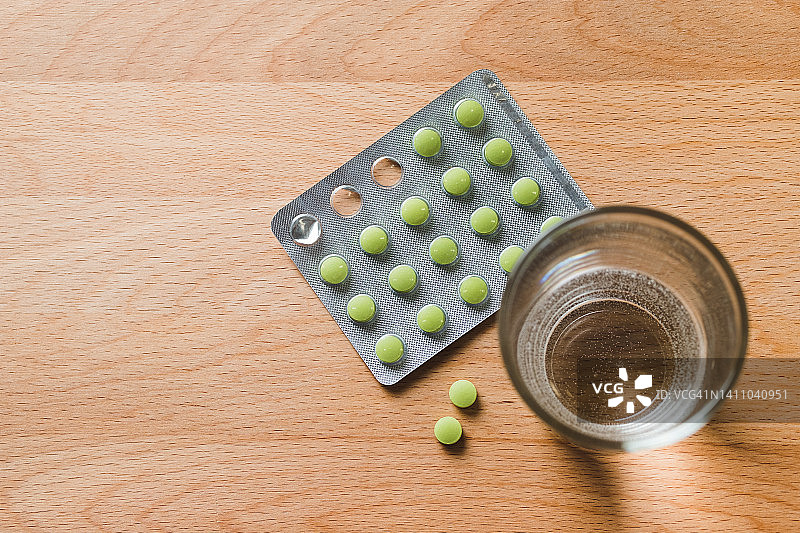 厨房的木桌上放着绿色药丸和一杯水。服用维生素、止痛药、药物和膳食补充剂。疾病预防和治疗的概念，保健和医药。为怀孕做计划和准备。图片素材