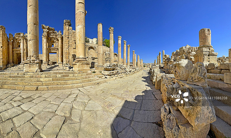 经典的科林斯式圆柱排列在杰拉什罗马遗址中心的石砌街道或车行道上。前景是复杂的装饰石雕。图片素材