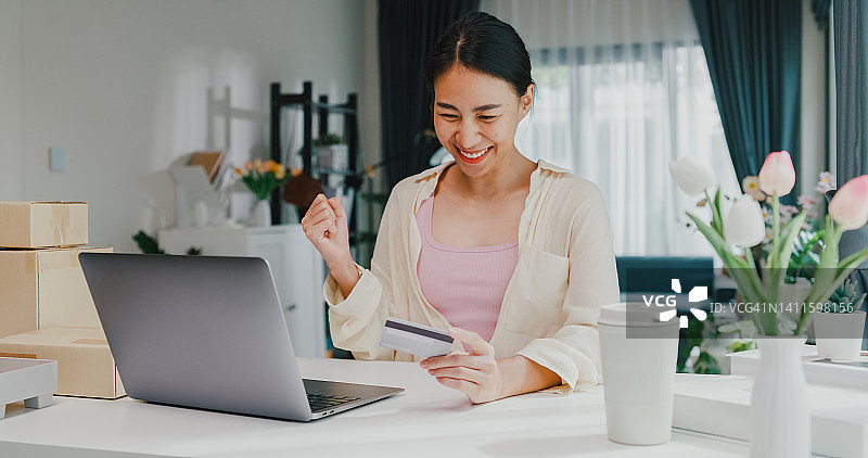 青年亚洲女女孩穿着休闲坐在书桌前与电脑笔记本电脑持有信用卡感到高兴和兴奋的成功付款在网上购物在客厅在家里。图片素材