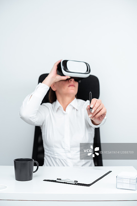 商业女性佩戴虚拟现实眼镜和学习专业技能通过未来模拟。现代训练中握笔坐在书桌前的女人。图片素材
