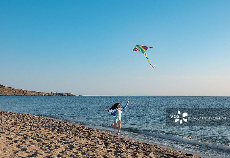 快乐的年轻女子在海滩上跑放风筝，在浅水中溅起水花。图片素材