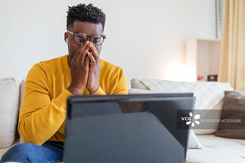 一个年轻人在家工作时看起来压力很大。沮丧的商人坐在家里的桌子上，用手托着自己的头。工作过度的年轻非洲裔美国商人坐在笔记本电脑前，抱着头。图片素材