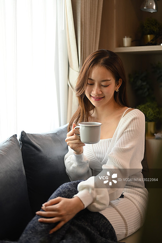 愉快的亚洲女人享受她的早晨咖啡，同时放松在家里的沙发上。休闲活动，积极情绪概念。图片素材