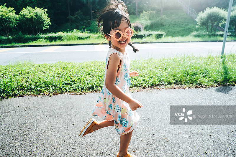 快乐的亚洲小女孩戴着花形太阳镜在公园里跑步。享受大自然和夏日户外。她对着镜头微笑。童年的生活方式，无忧无虑的自由理念图片素材