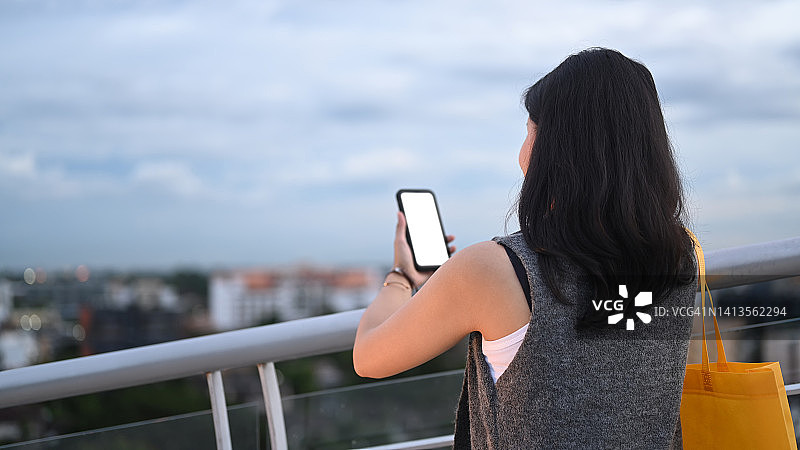 年轻女子使用手机的背影，站在露台上，背景是美丽的夜空。图片素材