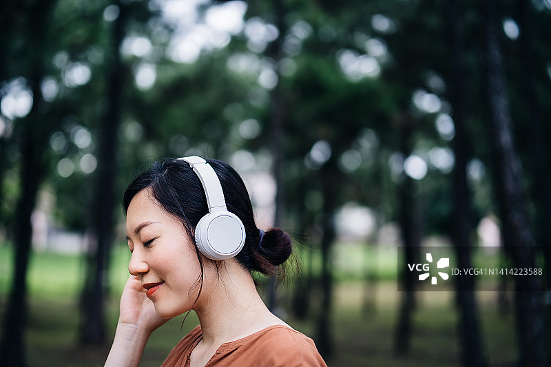 放松的年轻亚洲女子与眼睛闭上听音乐与耳机在自然，与树木周围她的户外。音乐、生活方式和科技图片素材