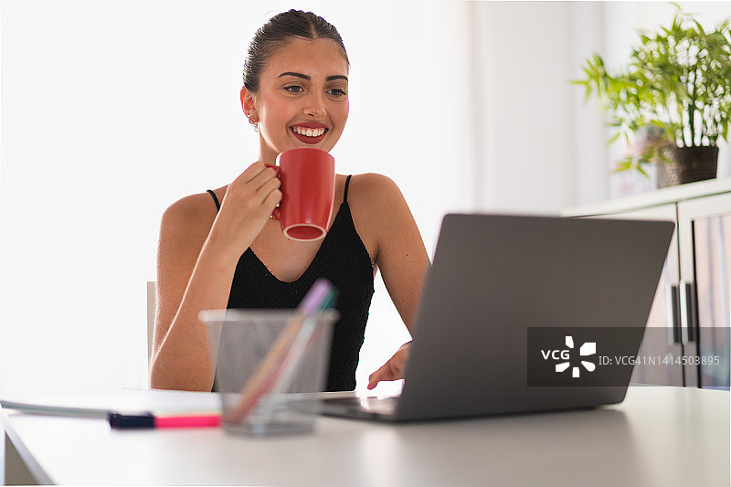 微笑的年轻女孩一边用笔记本电脑学习，一边喝着红杯子里的水图片素材