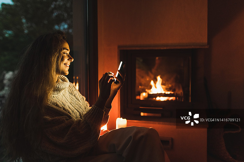 微笑的女人晚上坐在壁炉旁的窗台上，拿着手机图片素材