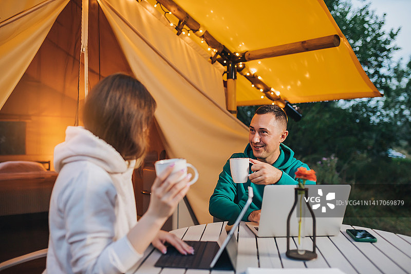 幸福的家庭夫妻自由职业者喝着咖啡，在夏天的晚上，在舒适的野营帐篷里用笔记本电脑工作。用于户外度假和度假的豪华露营帐篷。生活方式的概念图片素材