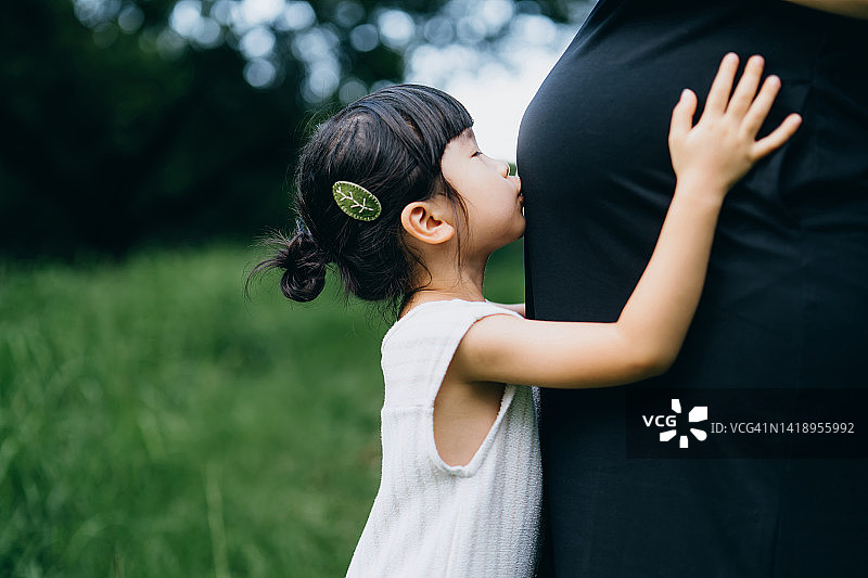 靠近可爱的亚洲小女孩拥抱和亲吻她怀孕的妈妈的肚子在自然。兄弟姐妹的爱。欢迎新的家庭成员。用爱和关怀期待新生活。分享爱的概念图片素材