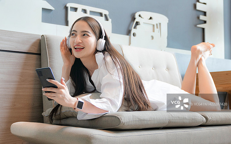 美丽的年轻女子戴着耳机在沙发上放松。女性用智能手机听音乐。放松和休闲的概念图片素材