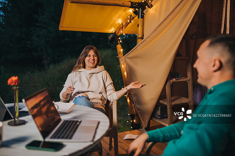 快乐的家庭夫妇自由职业者在舒适的野营帐篷里用笔记本电脑休息。户外度假度假的豪华露营帐篷。生活方式的概念图片素材